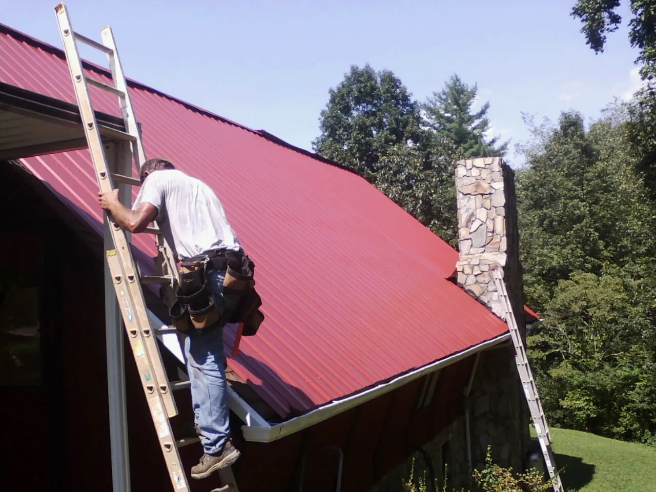 Roofer on a ladder beside a red metal roof and stone chimney — Soffit & Fascia Repair by Roofing Brothers in Summersville